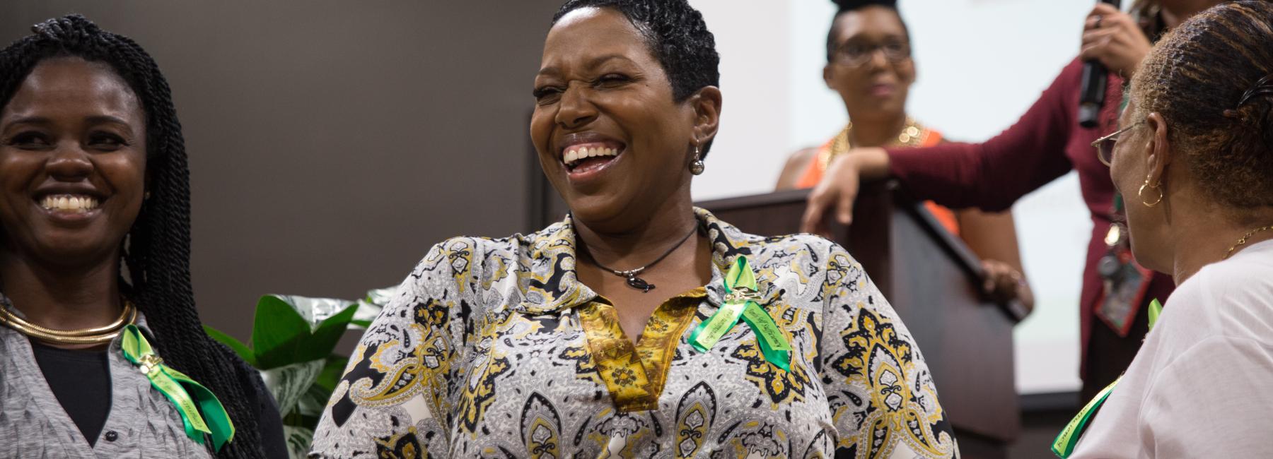 A group of women is gathered at an the LevelUp (formerly FSS) graduation. The woman in the center, wearing a patterned blouse, is laughing and appears to be the focus of the moment. She is joined by other smiling women, some of whom are wearing FSS Grad green ribbon pins. In the background, a woman stands at a podium, speaking into a microphone. The atmosphere is joyful and celebratory.